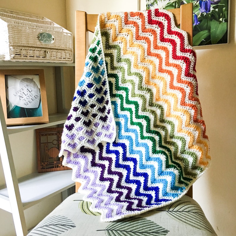 May include: A rainbow crochet baby blanket with a chevron pattern. The blanket is folded and draped over a wooden chair.