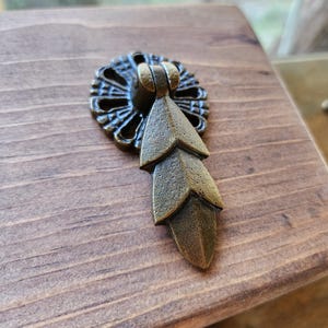 May include: Antique brass drawer pull with a leaf design. The pull is attached to a stained wooden surface. The pull has a decorative circular backplate and a three-leaf pendant. Ideal for furniture restoration or adding a vintage touch.