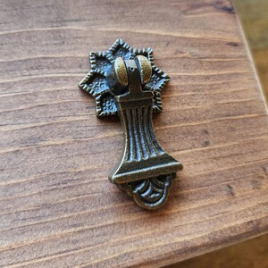 May include: A bronze-colored drawer pull with a starburst backplate. The pull has a decorative, ornate design with a curved handle and a detailed base. The hardware is set against a wooden surface.