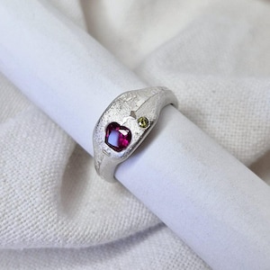 Może przedstawiać: Srebrny pierścionek z fakturowaną powierzchnią, ozdobiony różowym kamieniem szlachetnym w kształcie serca i mniejszym zielonym kamieniem szlachetnym.