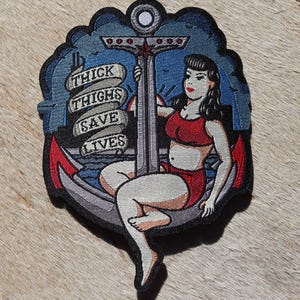 May include: A black and white embroidered patch with a pin-up girl sitting on an anchor. The anchor is surrounded by a blue background with white clouds. The pin-up girl is wearing a red bikini and has a red bow in her hair. The text "THICK THIGHS SAVE LIVES" is written on a banner wrapped around the anchor.