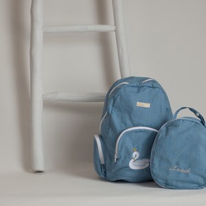 Könnte beinhalten: Zwei blaue Jeans-Rucksäcke mit weißem Besatz. Der größere Rucksack hat einen weißen Schwan mit einer goldenen Krone auf der Vorderseite gestickt. Der kleinere Rucksack hat das Wort "Leush" auf der Vorderseite gestickt.