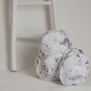 Könnte beinhalten: Zwei Rucksäcke mit Blumenmuster, einer größer als der andere, lehnen an einer weißen Holzleiter. Der größere Rucksack hat eine Reißverschlusstasche auf der Vorderseite. Beide Rucksäcke haben einen weißen Hintergrund mit einem grauen und weißen Blumenmuster.