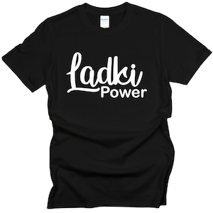 Può includere: T-shirt nera con la scritta bianca "Ladki Power" in corsivo. La parola "Power" è in un carattere più piccolo sotto "Ladki". La maglietta è in materiale morbido e ha uno scollo tondo.
