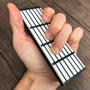 May include: A white and black guitar fretboard trainer held in a hand. The trainer has six black lines representing the strings and black bars. The fretboard is rectangular and appears to be made of plastic.