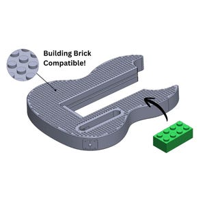 Könnte beinhalten: Eine graue, mit Bausteinen kompatible Gitarrenbasis mit einem grünen Baustein. Die Basis hat eine strukturierte Oberfläche mit runden Noppen zur Befestigung von Bausteinen. Der Text lautet "Building Brick Compatible!"