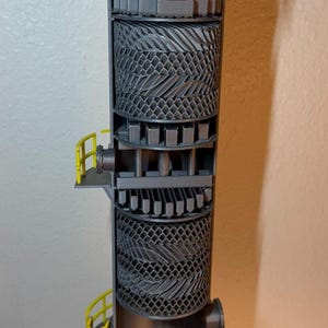 May include: A detailed model of an industrial distillation column, showing a cross-section. The model is mainly grey with yellow accents, including ladders and platforms. The internal components are visible, showcasing the column's structure.