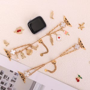 Puede incluir: Correa de reloj dorada con múltiples dijes, incluyendo una concha, una bota de vaquero, una carta de juego y un trébol de cuatro hojas. La correa está diseñada para un reloj inteligente y presenta un estilo de cadena.