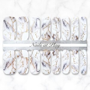 May include: Nail art strips with a white marble design featuring gold and gray veining. The strips are arranged on a silver backing with the text "Nails at Play".