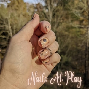 May include: A hand with a light pink nail polish base and sunflower nail decals. The text "Nails At Play" is written in pink cursive at the bottom of the image.