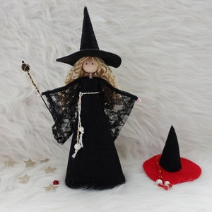 May include: A black felt witch doll with blonde hair, wearing a black dress with lace sleeves and a black pointed hat. The doll is holding a black and gold wand. The doll is standing on a white fluffy surface with gold stars scattered around it. There is a red felt witch hat with gold trim and white beads on the surface.
