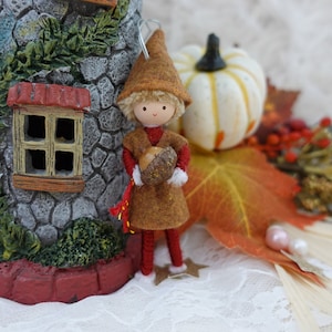 Peut inclure: Ornement sur le thème de l'automne avec une poupée miniature tenant un gland. La poupée porte une tenue en feutre marron et un chapeau pointu. Une petite maison en pierre avec un toit rouge et une citrouille blanche sont en arrière-plan.