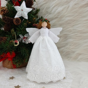 May include: White angel tree topper with a white lace dress, red berry wreath, and wings. The angel has blonde hair and a sweet expression.
