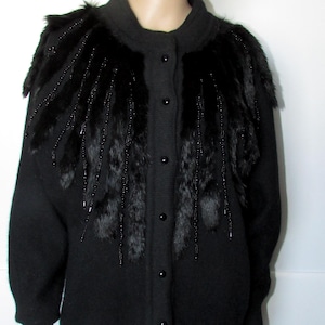 May include: A black long-sleeved cardigan featuring a round neckline and a front button closure. The upper chest area is decorated with black fur accents and dangling black beaded strands that extend downwards from the shoulder regions.