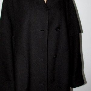 May include: A long, black coat with a wide collar and button-down closure. The coat has a classic design with a tailored fit, featuring a few buttons down the front and a simple, elegant style. The sleeves are long and the coat appears to be made of a heavy fabric.