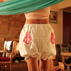 May include: White bloomers with red and white graphic patches. The bloomers have a ruffled hem and a gathered waist. The patches feature a red and white illustration of a person in a hat.