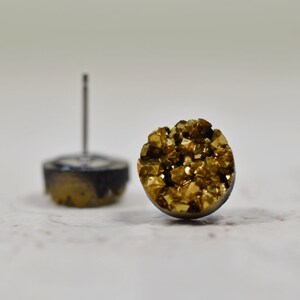 Könnte beinhalten: Ein Paar goldene und schwarze Ohrstecker mit rundem, strukturiertem Design. Die Ohrringe sind aus Metall gefertigt und haben eine glänzende Oberfläche.