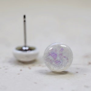 May include: A pair of white iridescent earrings with a silver post. The earrings have a round shape and a subtle purple shimmer.
