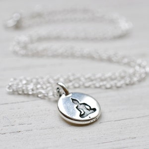 May include: A silver-colored necklace with a small, round pendant featuring a seated Buddha figure. The pendant and chain are displayed on a white wooden surface. The chain is delicate and the pendant has a detailed design.