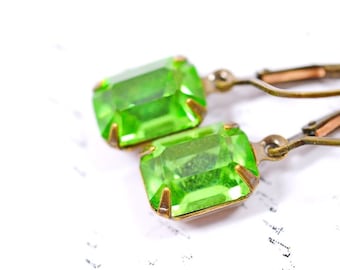 May include: A pair of leverback earrings featuring bright green, rectangular crystals. The crystals are set in a bronze-colored metal. The earrings are displayed on a white surface with faint patterns.