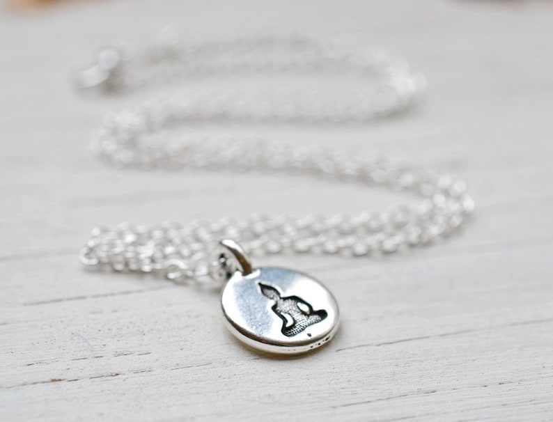 May include: A silver-colored necklace with a small, round pendant featuring a seated Buddha figure. The pendant and chain are displayed on a white wooden surface. The chain is delicate and the pendant has a detailed design.