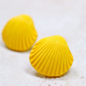 May include: Two bright yellow seashell-shaped objects. The objects have a ridged texture and a rounded shape, resembling small scallop shells. The background is a soft, neutral color, highlighting the vibrant yellow of the shells.