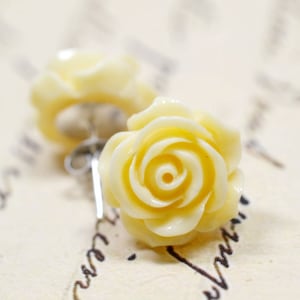 May include: Close-up of two pale yellow rose stud earrings. The earrings are made of a smooth material and feature detailed petals. The earrings are set against a background of handwritten script.