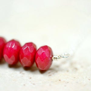 May include: Close-up of a necklace featuring faceted, deep red beads. The beads are strung together with a silver chain and wire. The background is a soft, neutral color, highlighting the vibrant red of the beads.