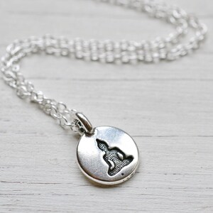 May include: A silver-colored necklace featuring a small, round pendant with a seated Buddha silhouette. The pendant hangs from a delicate chain, creating a minimalist and spiritual aesthetic. The necklace is suitable for everyday wear.