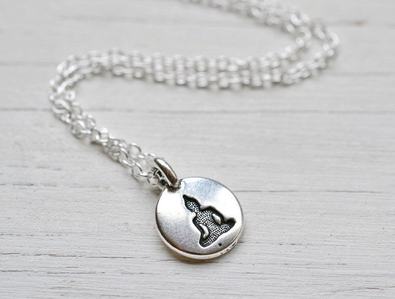 May include: A silver-colored necklace featuring a small, round pendant with a seated Buddha silhouette. The pendant hangs from a delicate chain, creating a minimalist and spiritual aesthetic. The necklace is suitable for everyday wear.