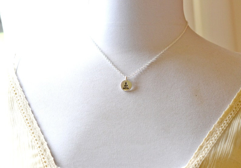 May include: A silver necklace featuring a small, round pendant with a seated Buddha figure. The pendant is attached to a delicate chain, displayed on a white mannequin. The necklace is a piece of jewelry.