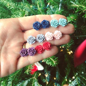 Puede incluir: Un conjunto de diez pendientes en forma de rosa de varios colores, incluyendo azul, rosa, rojo, morado y gris. Los pendientes se muestran en una mano sobre un fondo verde.