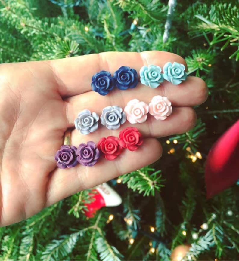 Puede incluir: Un conjunto de diez pendientes en forma de rosa de varios colores, incluyendo azul, rosa, rojo, morado y gris. Los pendientes se muestran en una mano sobre un fondo verde.