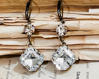 May include: A pair of dangle earrings with a square crystal and a smaller round crystal. The earrings are made with a brass finish and have a hook closure.