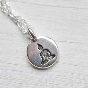 May include: A silver-colored pendant necklace featuring a small, round charm with a detailed cutout of a seated Buddha figure. The chain is a delicate, linked design.