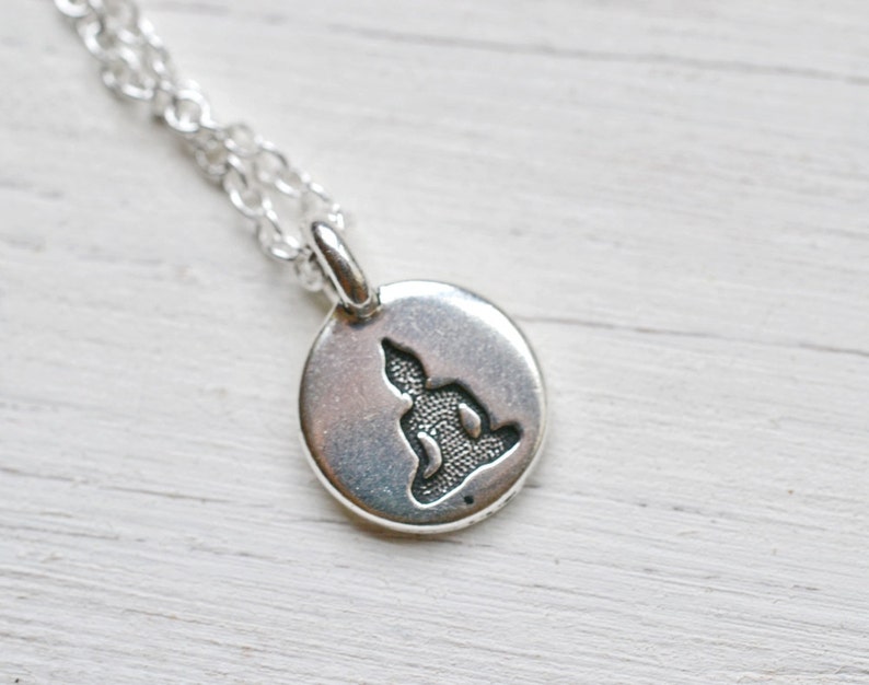 May include: A silver-colored pendant necklace featuring a small, round charm with a detailed cutout of a seated Buddha figure. The chain is a delicate, linked design.