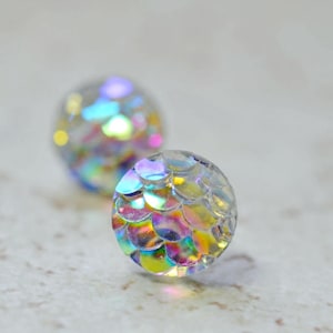 May include: Two clear, round stud earrings with a shimmering, iridescent, scale-like design. The earrings reflect a rainbow of colors, including pink, yellow, and blue. The earrings are set against a neutral background.