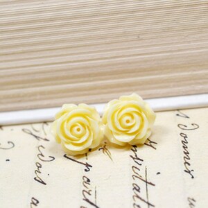 May include: Two pale yellow rose-shaped stud earrings. The earrings are set against a backdrop of vintage paper with handwritten script and a book. The earrings are a light, creamy yellow color and have detailed petals.