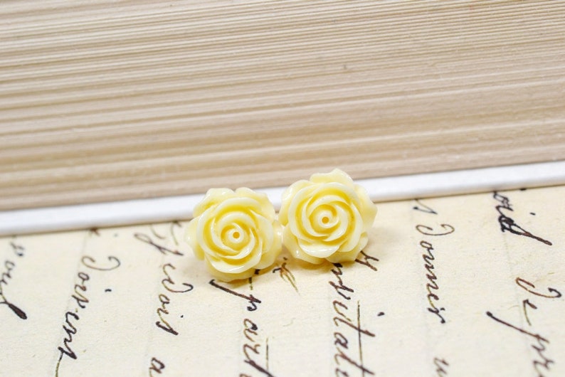 May include: Two pale yellow rose-shaped stud earrings. The earrings are set against a backdrop of vintage paper with handwritten script and a book. The earrings are a light, creamy yellow color and have detailed petals.