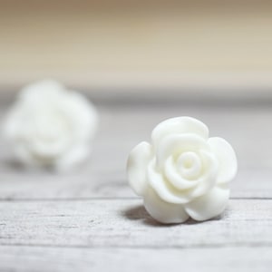 May include: Two white ceramic rose flower embellishments on a white wooden surface.