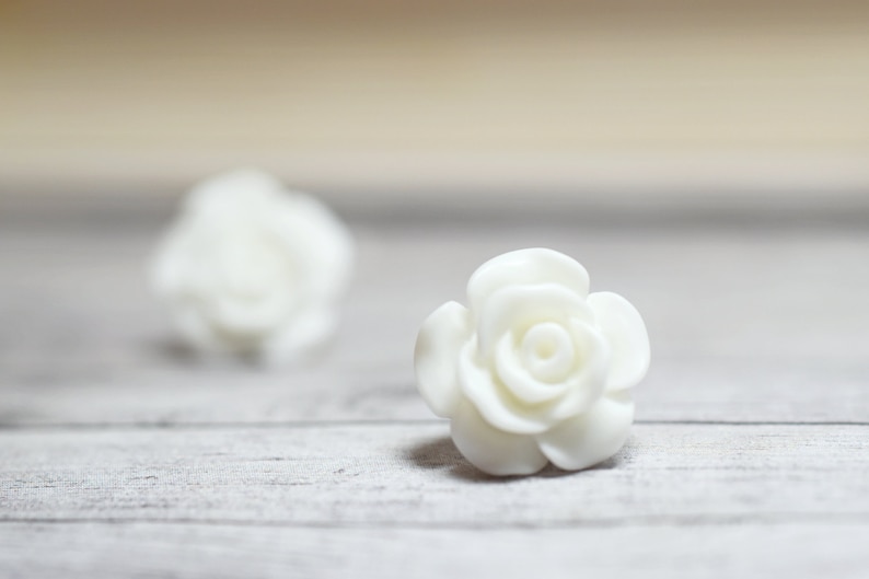 May include: Two white ceramic rose flower embellishments on a white wooden surface.