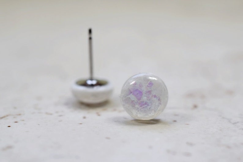 May include: A pair of white iridescent earrings with a silver post. The earrings have a round shape and a subtle purple shimmer.