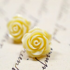 May include: Two pale yellow rose-shaped earrings. The earrings are in focus, with a soft, creamy color and detailed petal design. The background is a blurred, off-white surface with faint script.