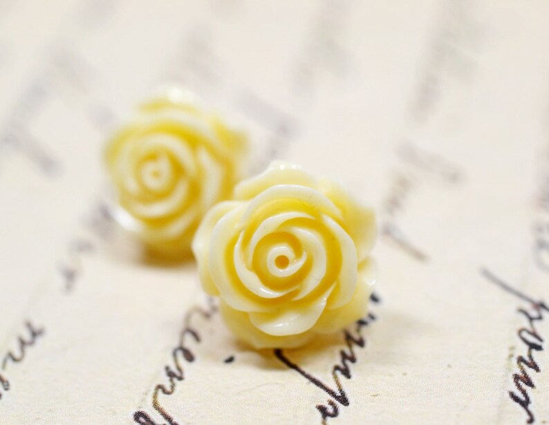 May include: Two pale yellow rose-shaped earrings. The earrings are in focus, with a soft, creamy color and detailed petal design. The background is a blurred, off-white surface with faint script.