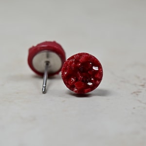 May include: A pair of red stud earrings with a textured, speckled finish. The earrings are made of a resin-like material and have a silver post.
