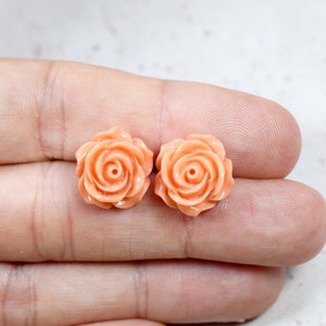 May include: Two peach-colored rose-shaped stud earrings. The earrings are made of a smooth material and feature detailed petals and a central spiral. The earrings are displayed on a light background.