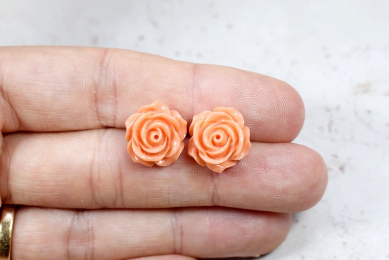 May include: Two peach-colored rose-shaped stud earrings. The earrings are made of a smooth material and feature detailed petals and a central spiral. The earrings are displayed on a light background.
