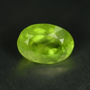 Op de afbeelding: Een gefacetteerde, ovaal geslepen peridot edelsteen. De edelsteen heeft een levendige, doorschijnende groene kleur, met zichtbare interne insluitsels. De steen is geplaatst tegen een donkere achtergrond, wat de helderheid en glans benadrukt. De slijping toont de facetten van de edelsteen.