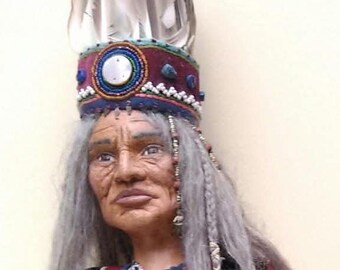 May include: A handcrafted doll with long gray hair wearing a maroon beaded headband featuring a central white circular inlay and upright gray feathers. The doll wears a black vest adorned with white buttons and intricate embroidery over a tan garment.