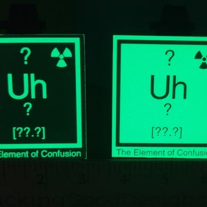 Può includere: Due adesivi quadrati verde brillante con la scritta "Uh" e punti interrogativi, che assomigliano a un elemento della tavola periodica. Il testo "The Element of Confusion" è in basso. È presente un simbolo di radiazione. Gli adesivi sono su sfondo scuro.